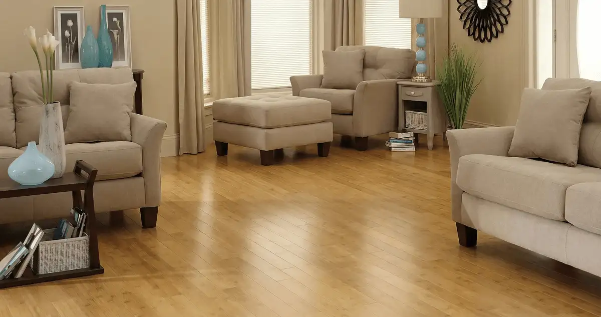 A living room with professionally installed bamboo flooring, beige sofas, a matching ottoman, a side table with a blue lamp, vases, framed pictures, and large windows with blinds and curtains in Hollywood, FL